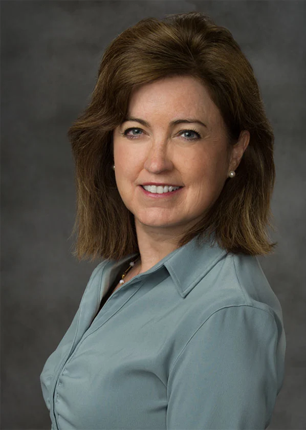 A photo of Eileen Maguire, who has shoulder-length dark brown hair and is wearing a blue-grey blouse.